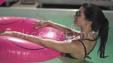 戴太阳镜的布鲁内特女士正拿着粉红色的橡胶圈在游泳池的水里。 穿比基尼的孤独女士休闲